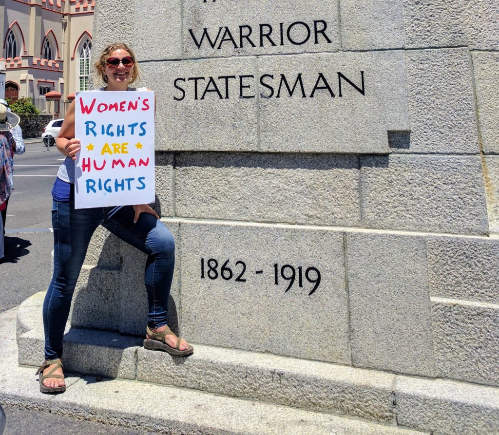 Julia holds a sign saying "Women's Rights are Human Rights" next to a statue podium reading "Warrior, Statesman"